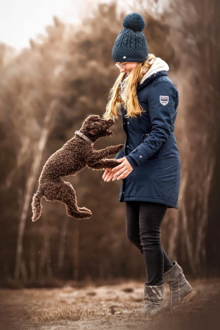 Eine junge Frau mit blauer Jacke und Mütze steht in der Natur und spielt mit ihrem braunen springenden Pudel.