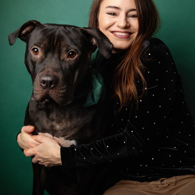 Eine hübsche Frau hält ihren schwarzen Pitbull im Arm vor einem dunkelgrünem Fotohintergrund.